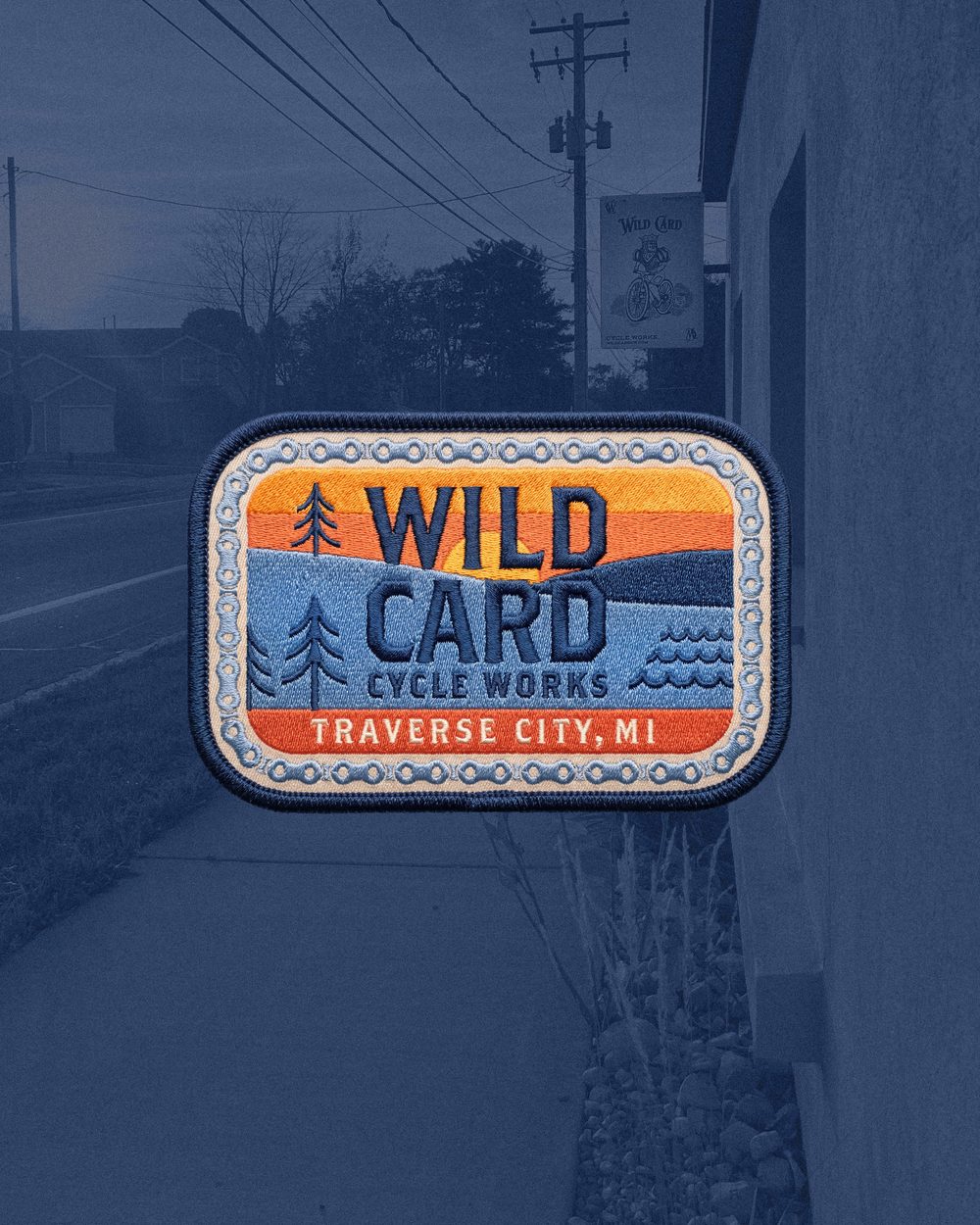 The Wild Card Cycle Works patch: rolling hills, conifers, and lake waves under a sunset, ringed by a bicycle chain, over a navy-tinted street scene