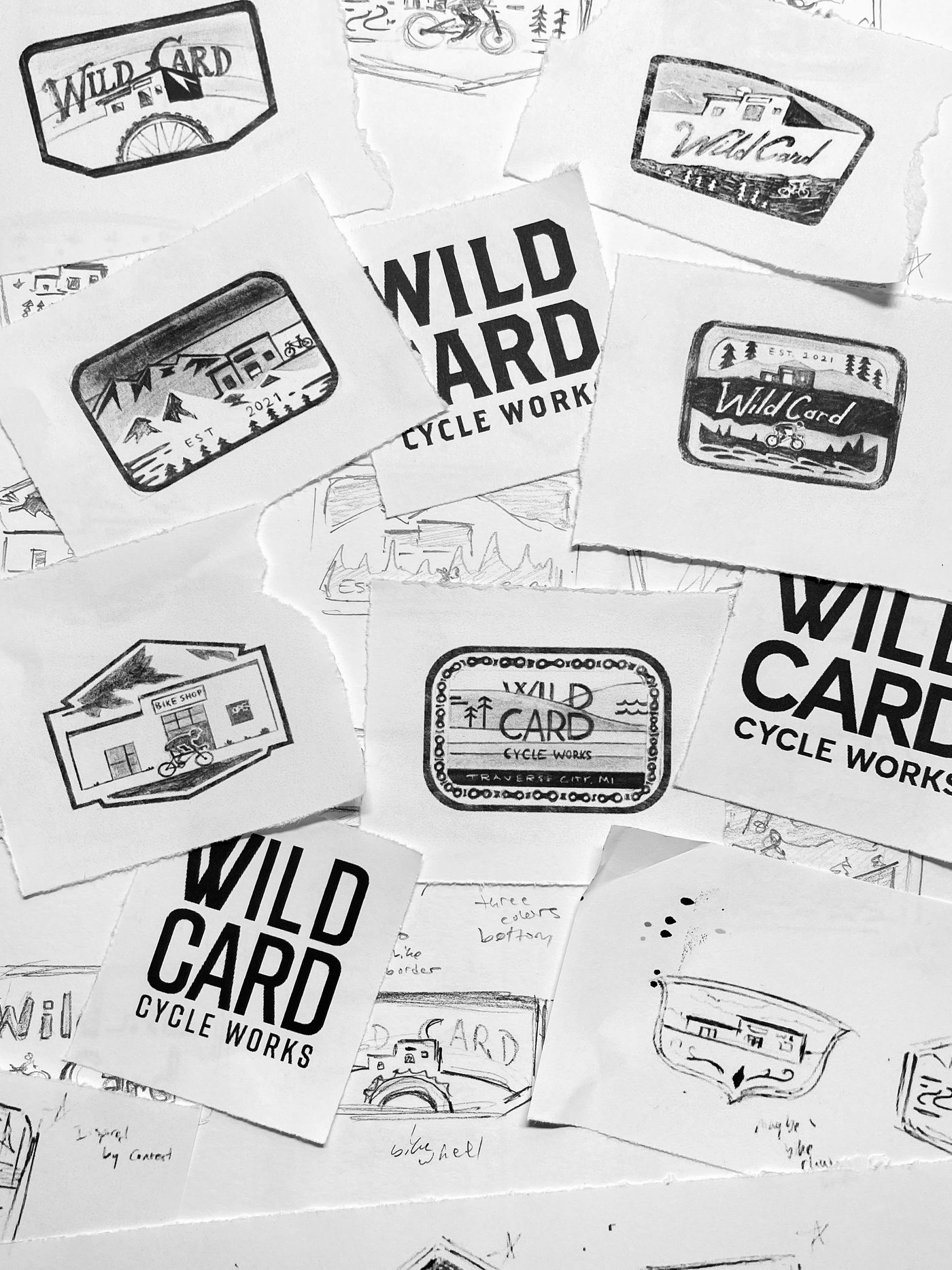 Pencil and marker sketches of Wild Card Cycle Works patch concepts spread across a table: badges, storefronts, and bold WILD CARD lettering